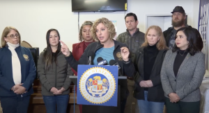 at a press conference. A woman with Assemblywoman Pilar Schivao is at a podium labeled "California State Assembly," with microphones from news outlets including FOX 11. She gestures with her hands while others behind her listen attentively.